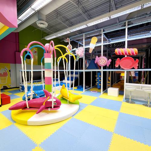 Colorful indoor play area featuring candy-themed decor, swings, and soft flooring in bright pastel colors.
