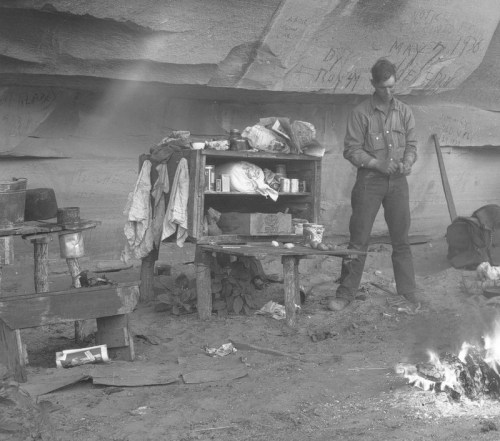 A man stands near a campfire in front of a rustic table, surrounded by personal items and a rocky backdrop.