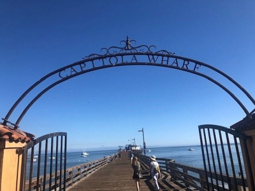 A sunny day at Capitola Wharf, with people walking along the wooden pier and boats in the calm water.