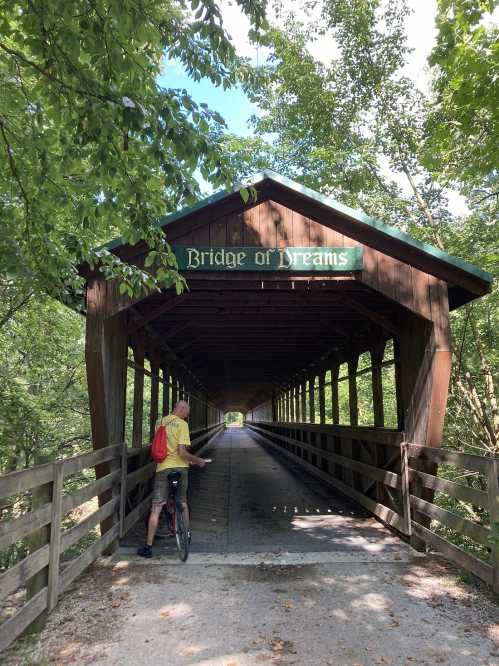 A cyclist approaches the "Bridge of Dreams," a wooden covered bridge surrounded by lush greenery.