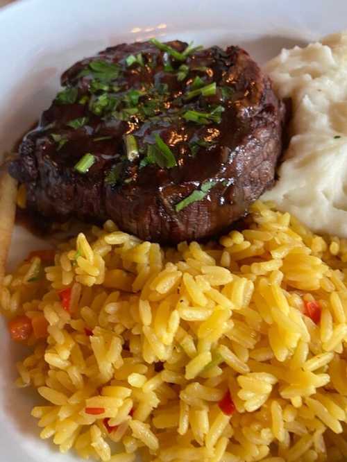 A plate featuring a grilled steak topped with herbs, served with yellow rice and creamy mashed potatoes.