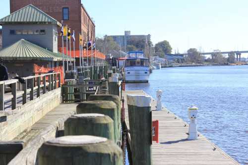 A peaceful waterfront scene with a dock, boats, and buildings along the river under a clear blue sky.