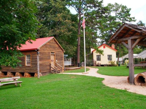 A scenic view of two historic wooden buildings with a flagpole, surrounded by green grass and trees.