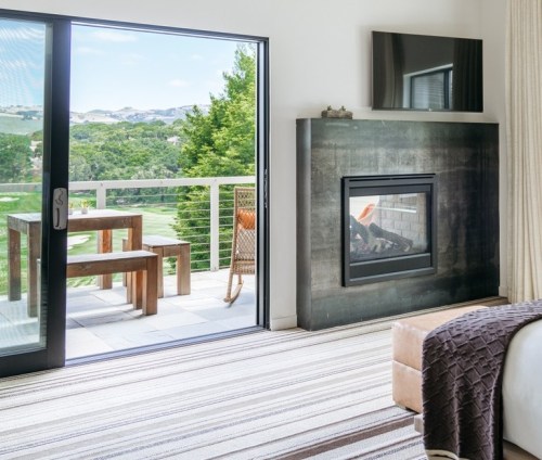 A modern bedroom with a fireplace, large window, and a view of a green landscape and outdoor seating area.