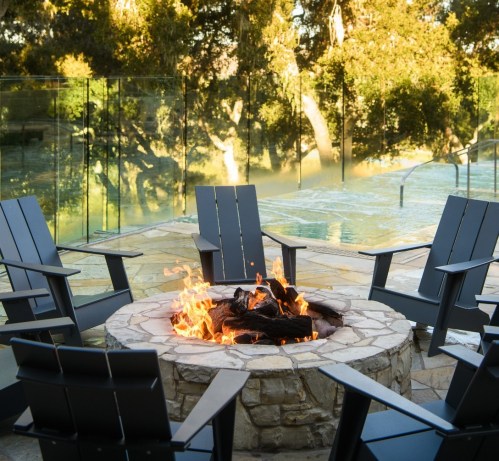 A cozy outdoor fire pit surrounded by black chairs, with a pool and trees in the background.