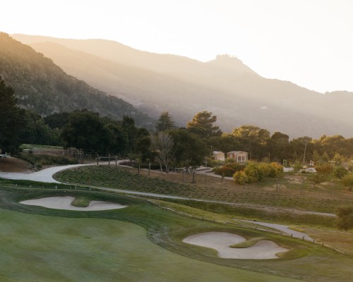A serene golf course at sunset, surrounded by rolling hills and trees, with sand traps visible on the green.