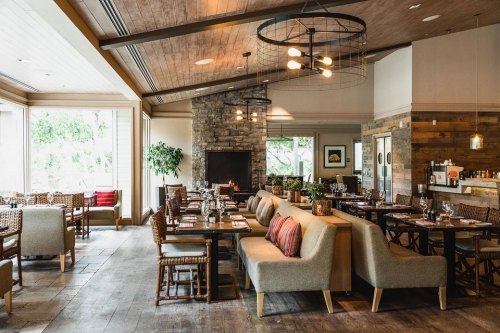 Cozy restaurant interior with wooden accents, comfortable seating, and a stone fireplace, featuring large windows and warm lighting.