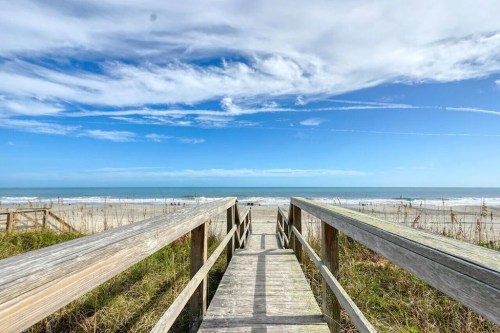 A wooden walkway leads to a sandy beach under a blue sky with scattered clouds.