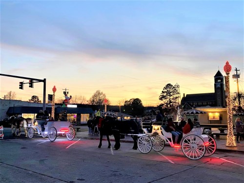 Carriage rides with festive lights in a town square at sunset, surrounded by holiday decorations and trees.