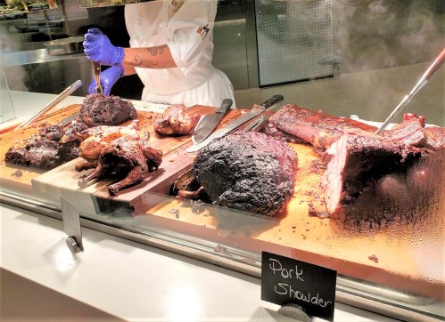 A chef in gloves carves various smoked meats, including pork shoulder, displayed on a wooden counter.