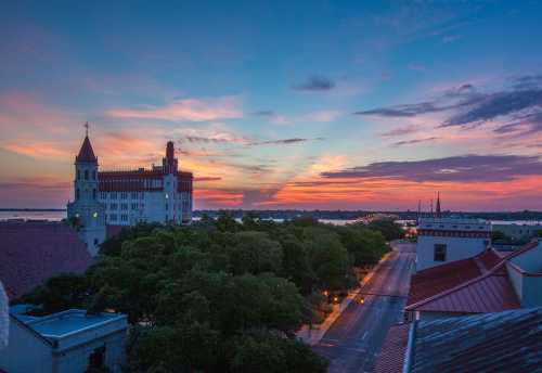 Sunset over a cityscape featuring a historic building, trees, and a calm waterway in the background.