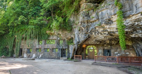 A stone building built into a rocky cliff, surrounded by lush greenery and featuring large windows and a wooden deck.