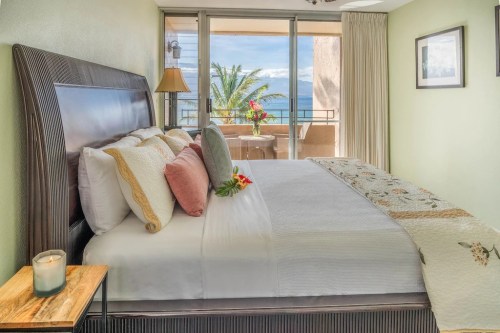 Cozy bedroom with a king-sized bed, decorative pillows, and a view of palm trees and ocean through large windows.