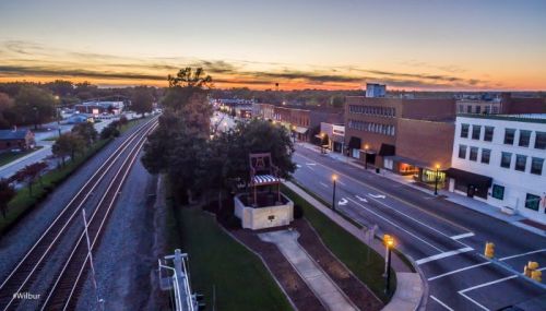 Aerial view of a small town at sunset, featuring train tracks, buildings, and a tree-lined street.