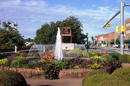 A decorative fountain surrounded by colorful flowers and greenery in a small town setting with buildings in the background.