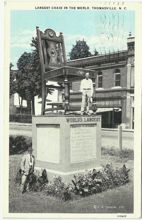 A man stands beside a giant chair monument labeled "World's Largest" in Thomasville, N.C., with buildings in the background.