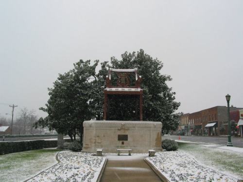 A snow-covered park with a large decorative arch and a tree, surrounded by a small garden and buildings in the background.