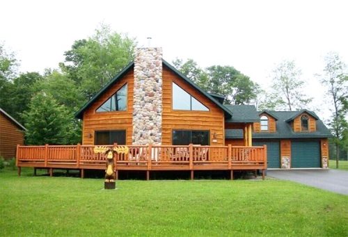 A large wooden house with a stone chimney, surrounded by greenery and featuring a spacious deck.