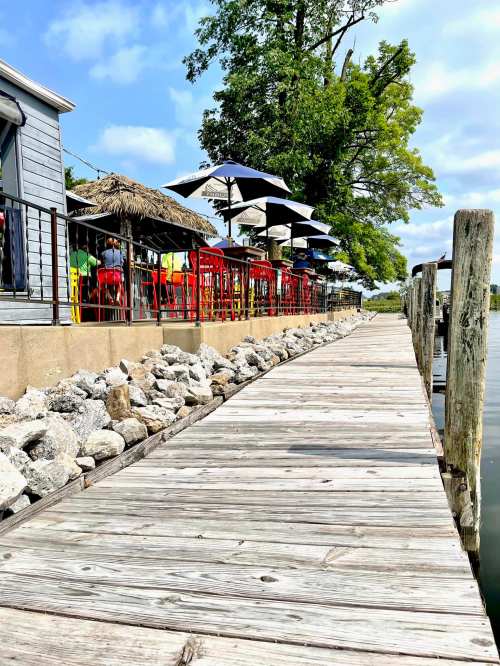 A wooden dock lined with umbrellas and colorful chairs, leading to a waterfront restaurant under a blue sky.
