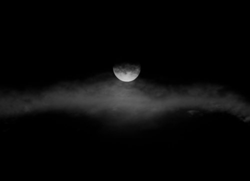 A partially obscured moon shines through dark clouds in a night sky.