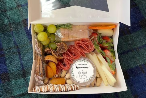 A charcuterie box with meats, cheeses, fruits, nuts, and a small jar, arranged neatly in a white box.
