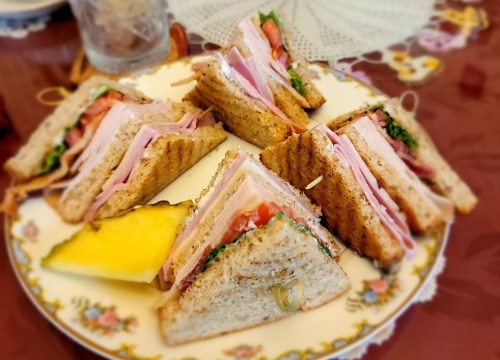 A plate of stacked sandwiches with ham, lettuce, and tomato, accompanied by a slice of pineapple.