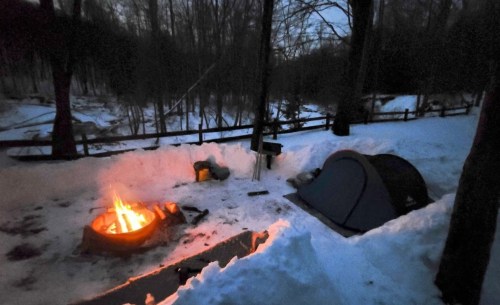 A campsite in the snow at dusk, featuring a glowing campfire and a tent surrounded by snowbanks and trees.
