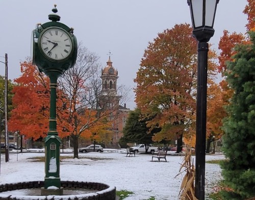 A green clock stands in a park with autumn trees and a dusting of snow, featuring a church tower in the background.