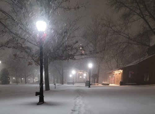 A snowy scene at night with streetlights illuminating a path and a building in the background, surrounded by falling snow.