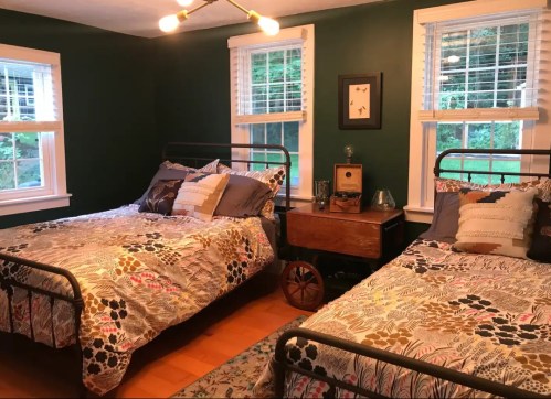 Cozy bedroom with two metal-framed beds, colorful bedding, and a wooden nightstand, surrounded by green walls and windows.