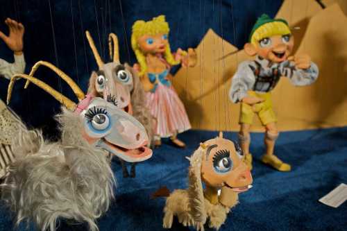 Colorful marionette puppets, including goats and a boy, displayed against a blue backdrop with mountains.