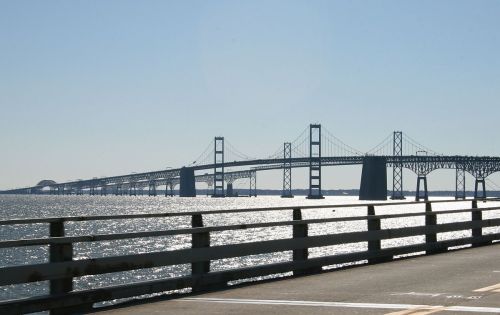 A long bridge spans across shimmering water under a clear blue sky, with a roadway in the foreground.