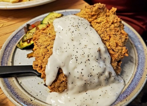 A plate of fried chicken topped with creamy white gravy and black pepper, served with sautéed vegetables on the side.