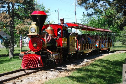 A colorful miniature train with passengers rides along a track in a park, surrounded by trees and greenery.