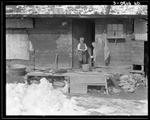 Two boys stand at the entrance of a rustic wooden cabin, surrounded by snow and scattered tools.