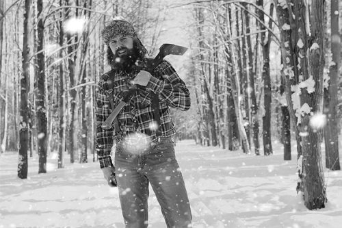 A man in a plaid shirt and hat holds an axe, standing in a snowy forest with falling snowflakes.