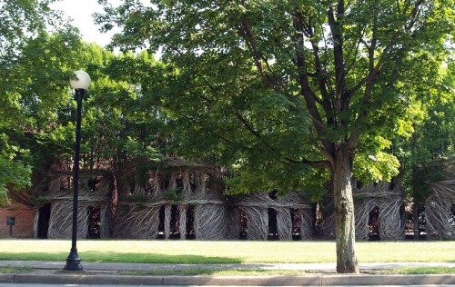 A large, intricate structure made of woven branches, surrounded by trees and grass, near a street lamp.