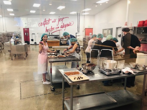 A kitchen scene with people preparing food, wearing aprons and hairnets, with a wall that says "crafted with joy."