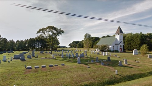 A peaceful cemetery with various gravestones and a small church in the background, surrounded by trees and grass.