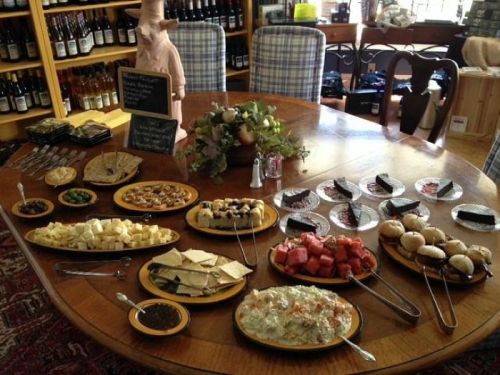 A table set with various cheeses, fruits, crackers, and desserts, surrounded by wine bottles in a cozy setting.