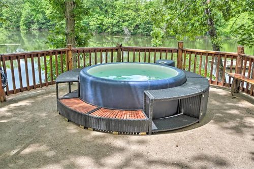 A hot tub on a wooden deck overlooking a serene lake surrounded by lush greenery.