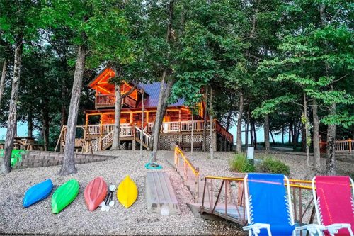 A log cabin surrounded by trees, with colorful kayaks and lounge chairs on a pebbled shore by a lake.