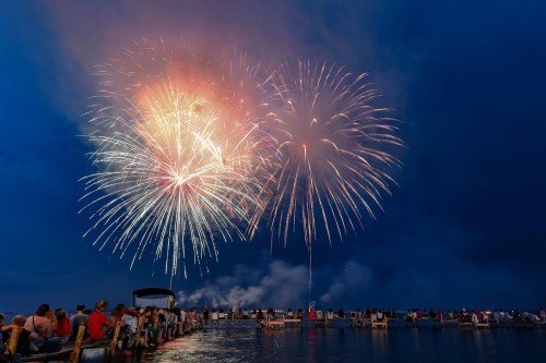 Colorful fireworks burst over a crowd gathered on a dock by the water, under a darkening sky.