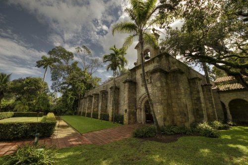 Historic stone building surrounded by lush greenery and palm trees under a cloudy sky.