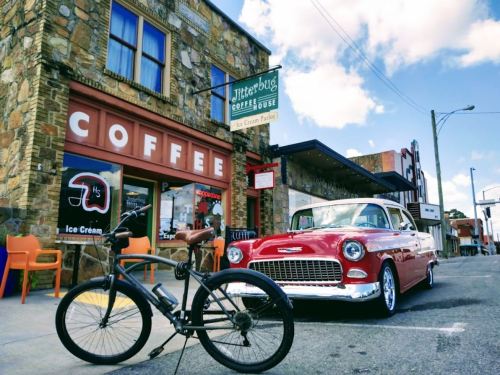 A vintage red car parked outside a coffee shop, with a bicycle in the foreground and a sunny sky above.