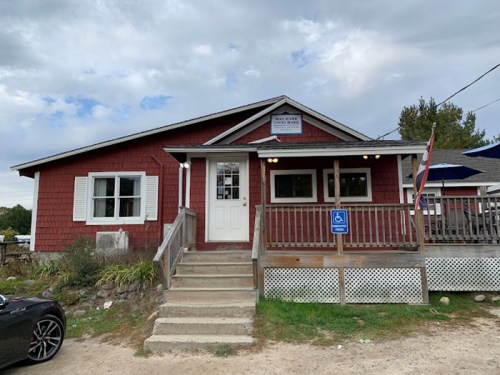 A red wooden building with a white door and steps, featuring a sign and accessible parking in front.