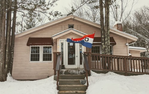 A cozy house with a wooden porch, surrounded by snow and trees, featuring a flag waving in the winter breeze.
