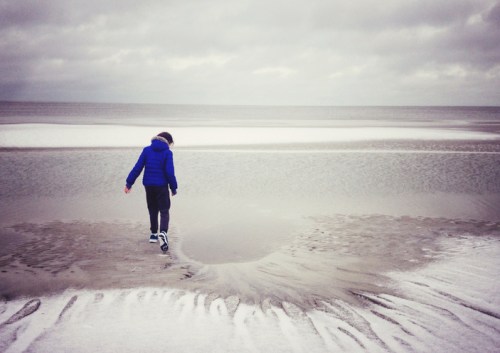 A person in a blue jacket walks along a sandy beach, with calm waters and overcast skies in the background.