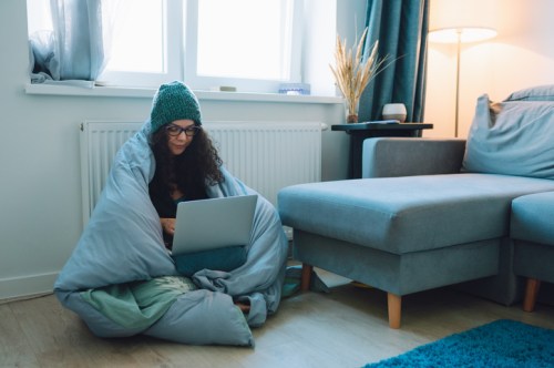 A woman sits on the floor wrapped in a blanket, using a laptop in a cozy, well-lit living room.