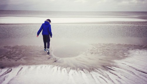 A child in a blue jacket walks along a sandy beach, with calm waves and a cloudy sky in the background.